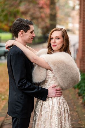 Newlywed couple embracing outdoors in fall, woman in champagne lace dress with white fur stole, man in black suit on tree-lined sidewalk.