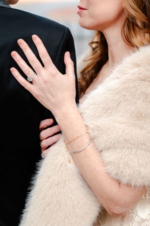 Bride in fur stole displays diamond engagement ring and gold bracelets while holding groom in black tuxedo.