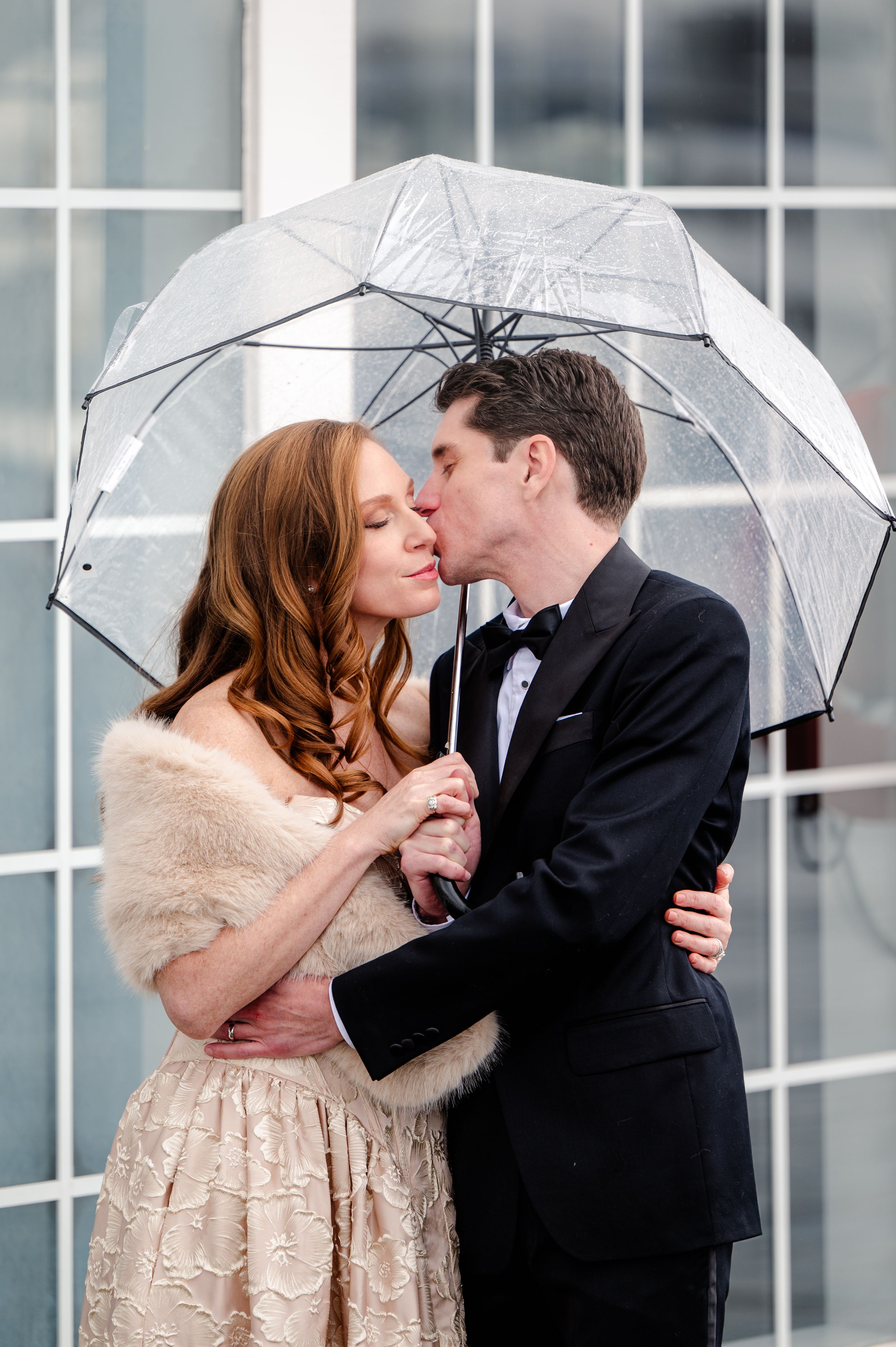 Romantic newlywed couple sharing a kiss under a clear umbrella in the rain, woman in fur stole and lace dress, man in black tuxedo.