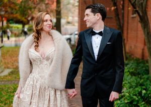 Bride in lace gown with fur stole holds hands with groom in black tuxedo, walking outdoors in autumn setting.