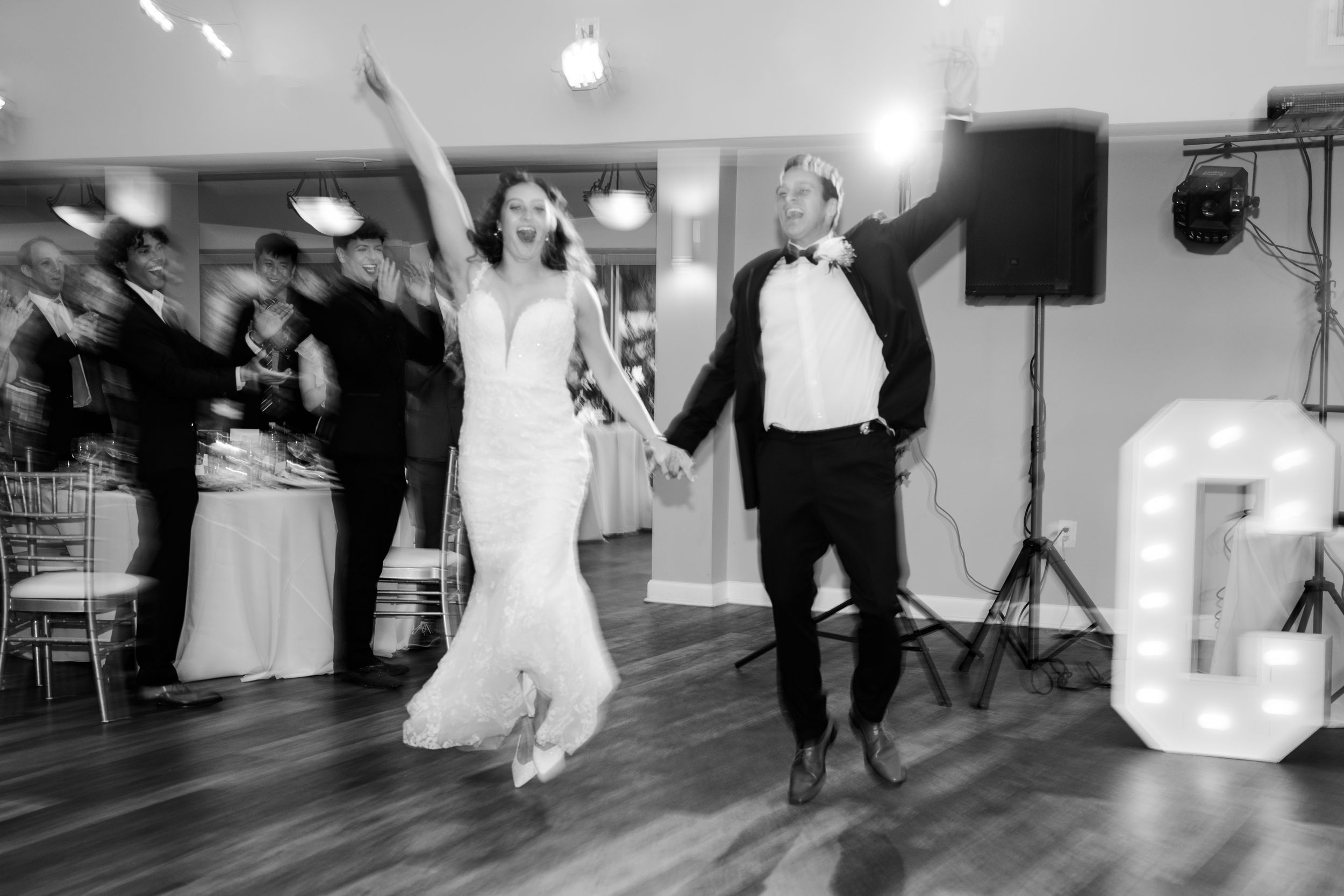 Black and white photo of bride and groom making grand entrance at wedding reception, arms raised in celebration.