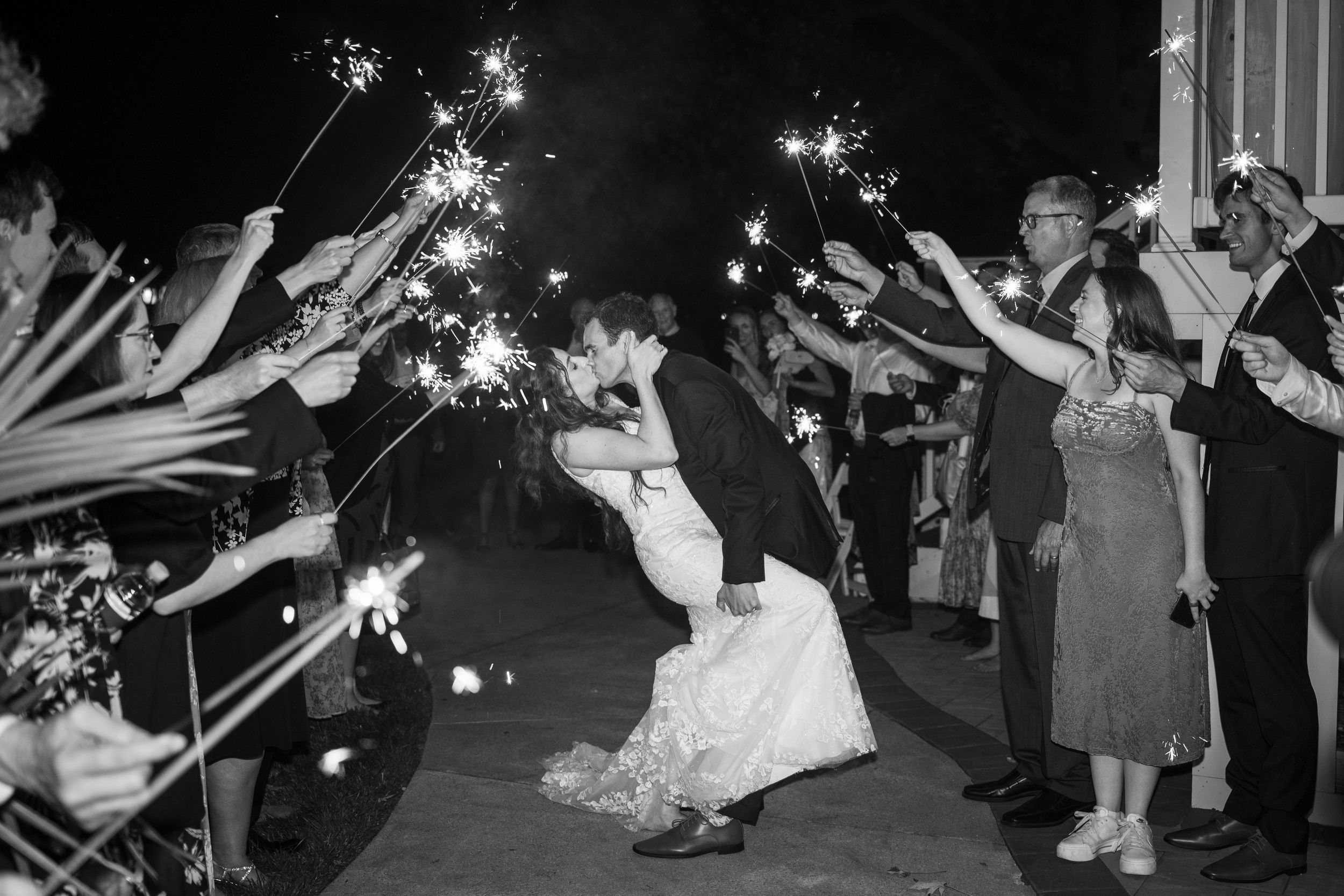 Black and white photo of newlyweds kissing under sparklers held by wedding guests during grand exit in Baltimore.