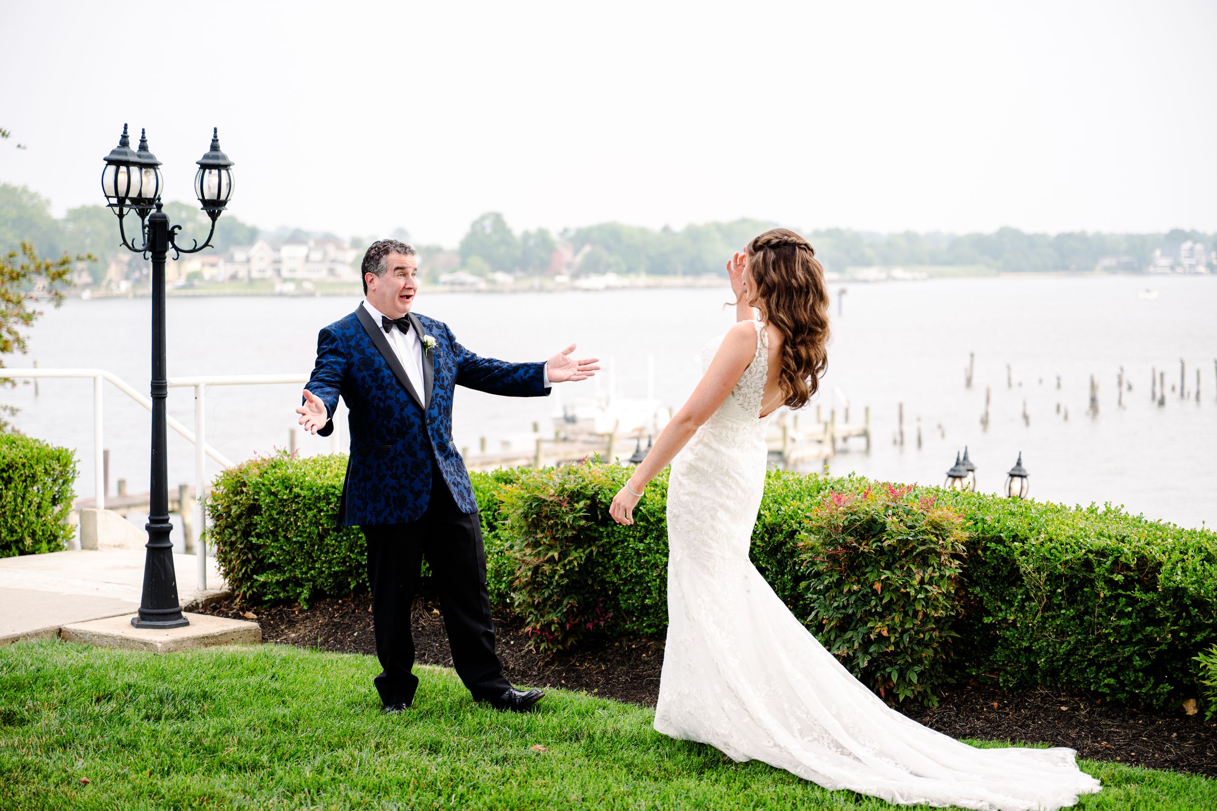 Bride in white gown and father of the bride in navy tuxedo share a joyful first look moment on a waterfront lawn in Baltimore.