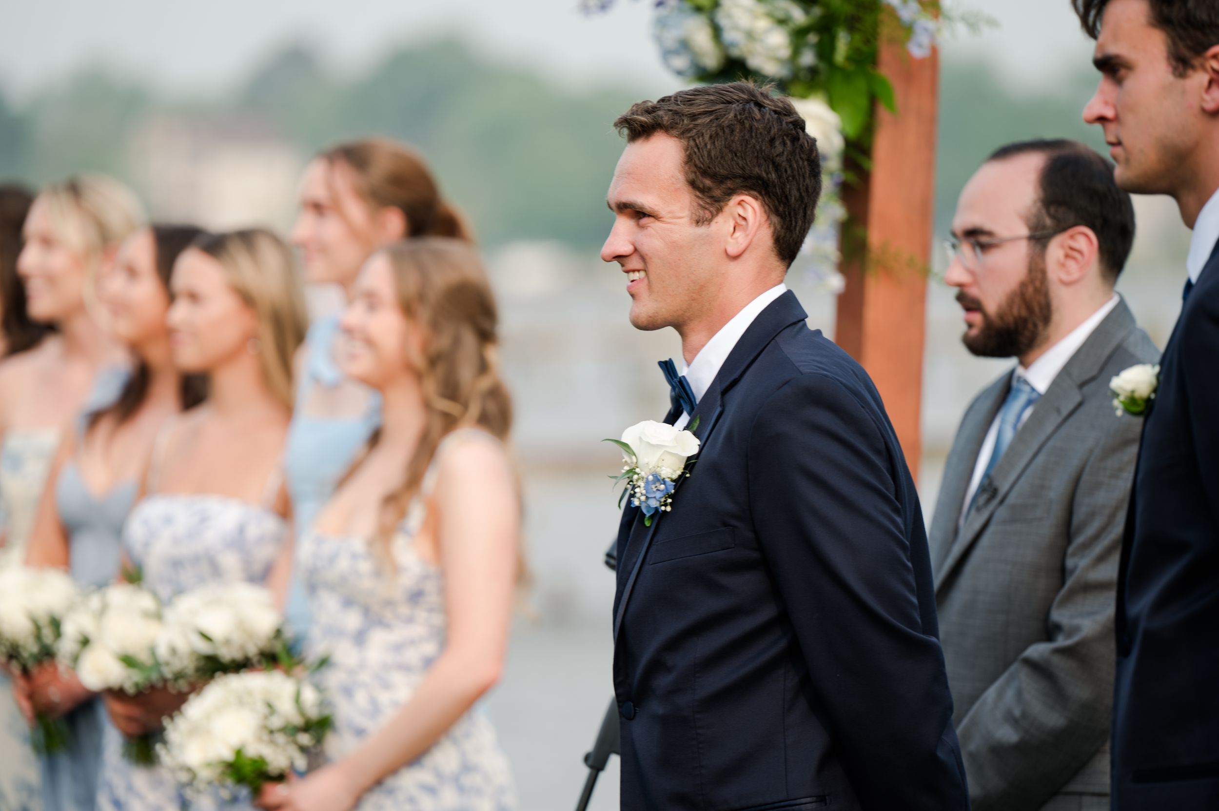 Groom in navy suit smiling at wedding ceremony with bridesmaids in blue dresses holding white floral bouquets in Baltimore.