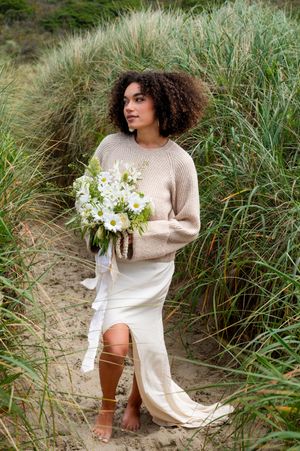 A bohemian bride in a cream sweater and dress holding white flowers amid tall beach grass.