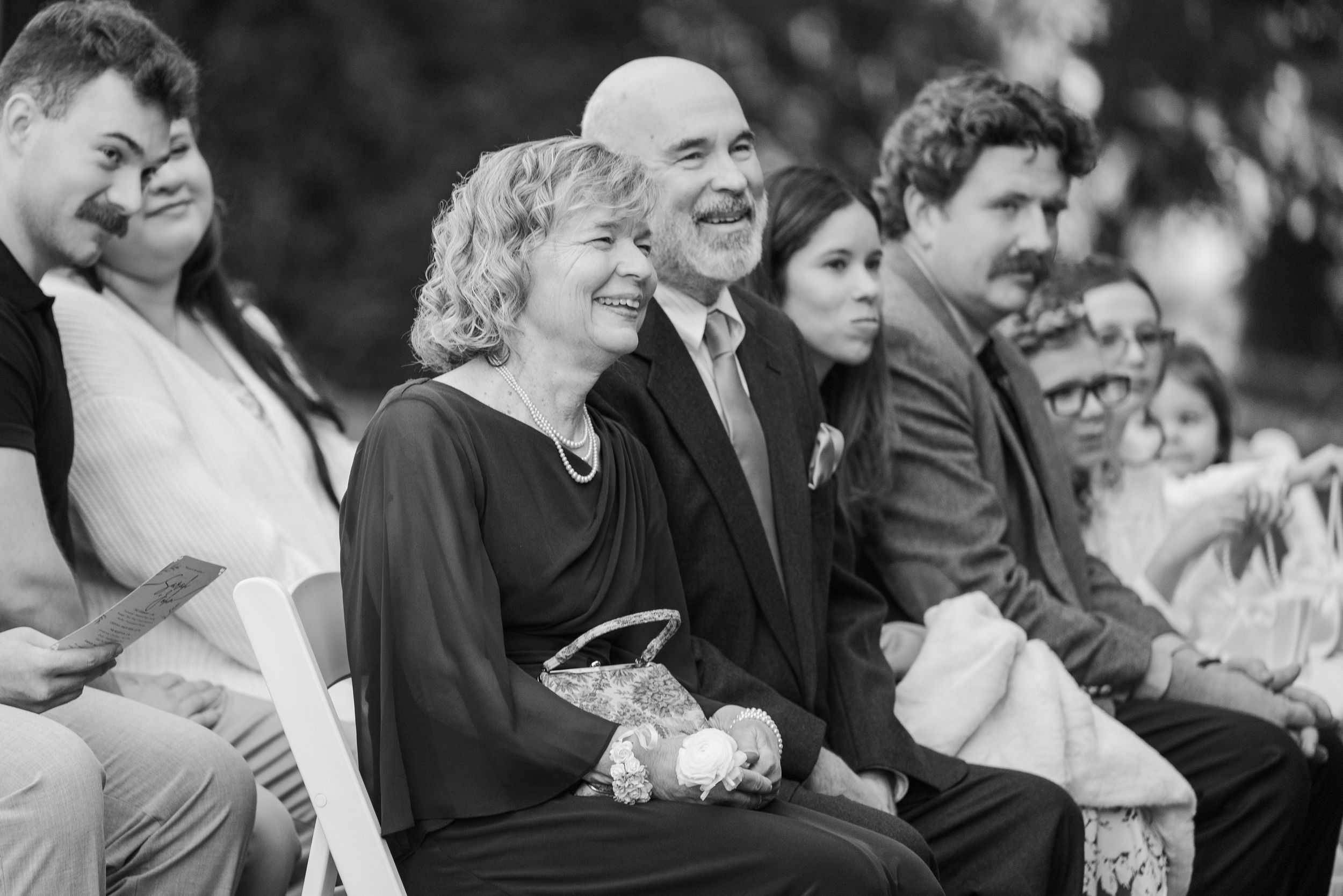 Smiling parents seated among wedding guests during outdoor ceremony, captured in classic black and white photography.