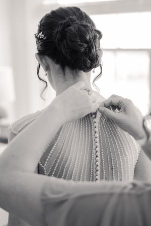 Bride getting pearl button wedding dress fastened from behind, showing elegant updo hairstyle with decorative pin.
