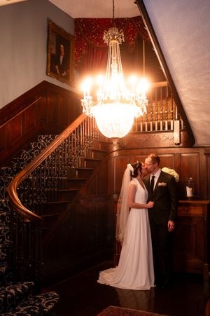 Bride and groom share a romantic kiss under a crystal chandelier in an elegant wood-paneled mansion staircase.