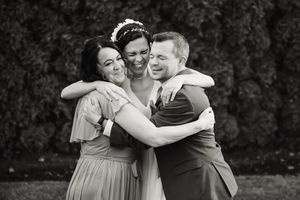 Black and white photo of a bride sharing a joyful group hug with a woman and man outdoors.