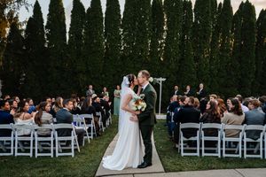 Outdoor wedding ceremony with bride and groom kissing at the altar, guests seated in white chairs, tall green trees backdrop.