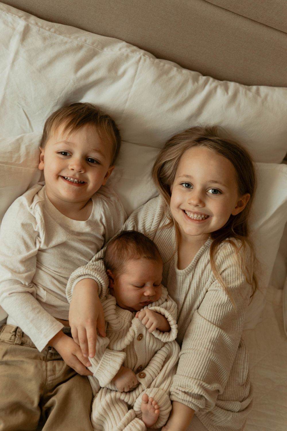 Siblings share sweet moments together while lying on cream colored bedding.
