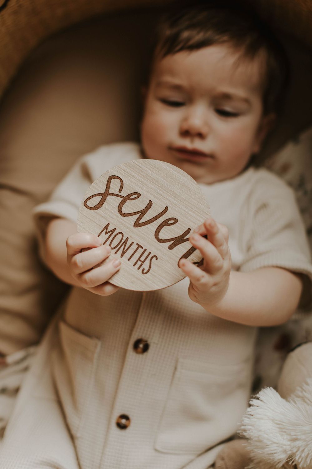 Seven month old baby holding a wooden milestone sign reading seven months, dressed in a cream outfit.