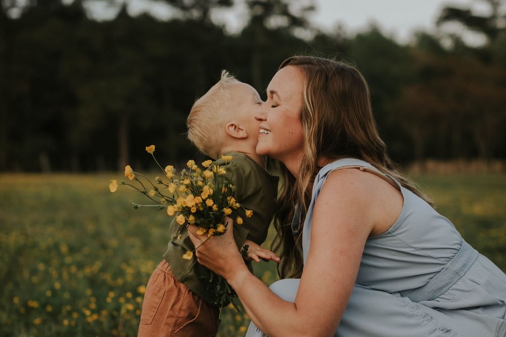 A mother and young child share a tender kiss in a sunny yellow flower field, with the child holding a bouquet of wildflowers.