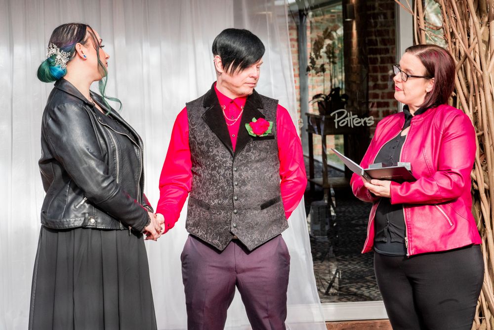 Bride in black dress and groom in black vest participate in an outdoor wedding ceremony with brick wall backdrop.
