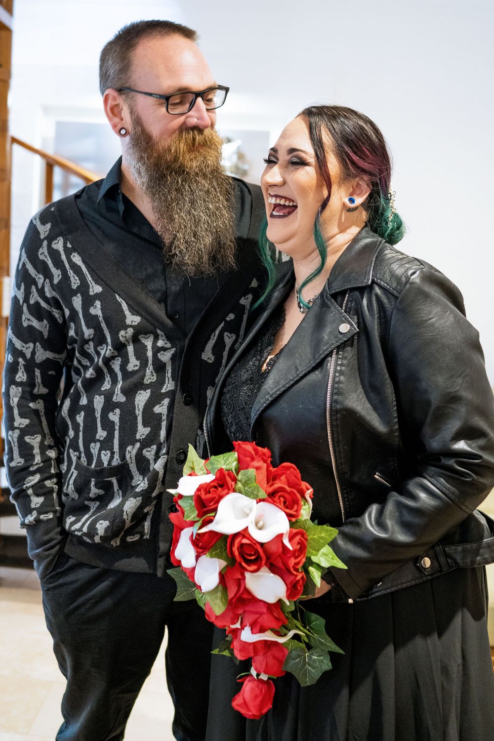 Alternative wedding couple sharing a joyful moment with red rose bouquet.