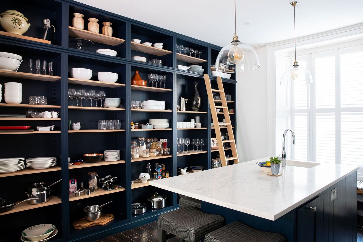 Floor-to-ceiling navy shelving unit stores kitchen items with rolling ladder for accessibility.