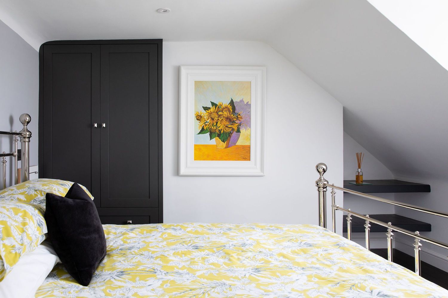 Attic bedroom with black wardrobe, yellow floral bedding, and framed artwork against white walls.
