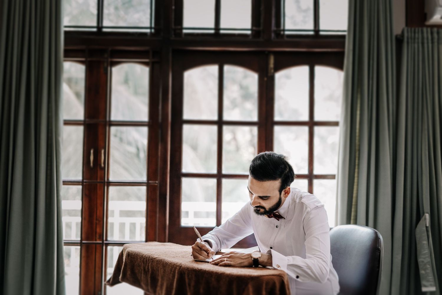 A person in a white shirt writes at a wooden desk near a large window with green curtains.