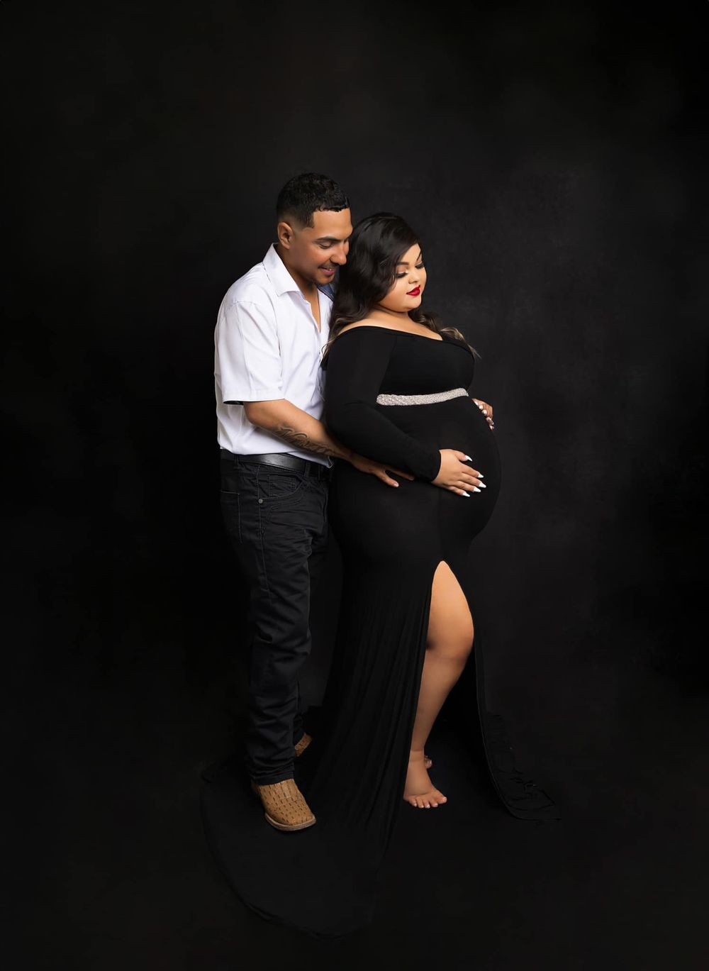 Expectant couple poses together in black and white outfits against dark backdrop.