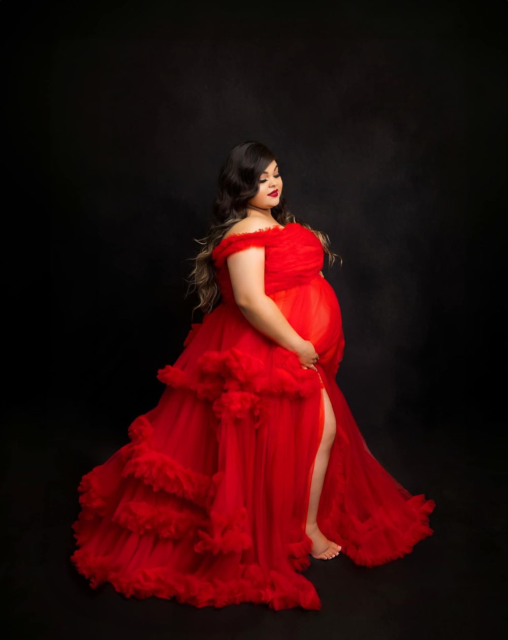 A glamorous red tulle maternity dress with ruffle details photographed against black.