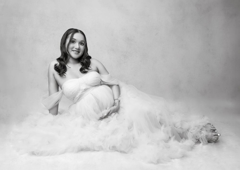 A serene black and white maternity portrait with the subject seated among billowing white fabric.