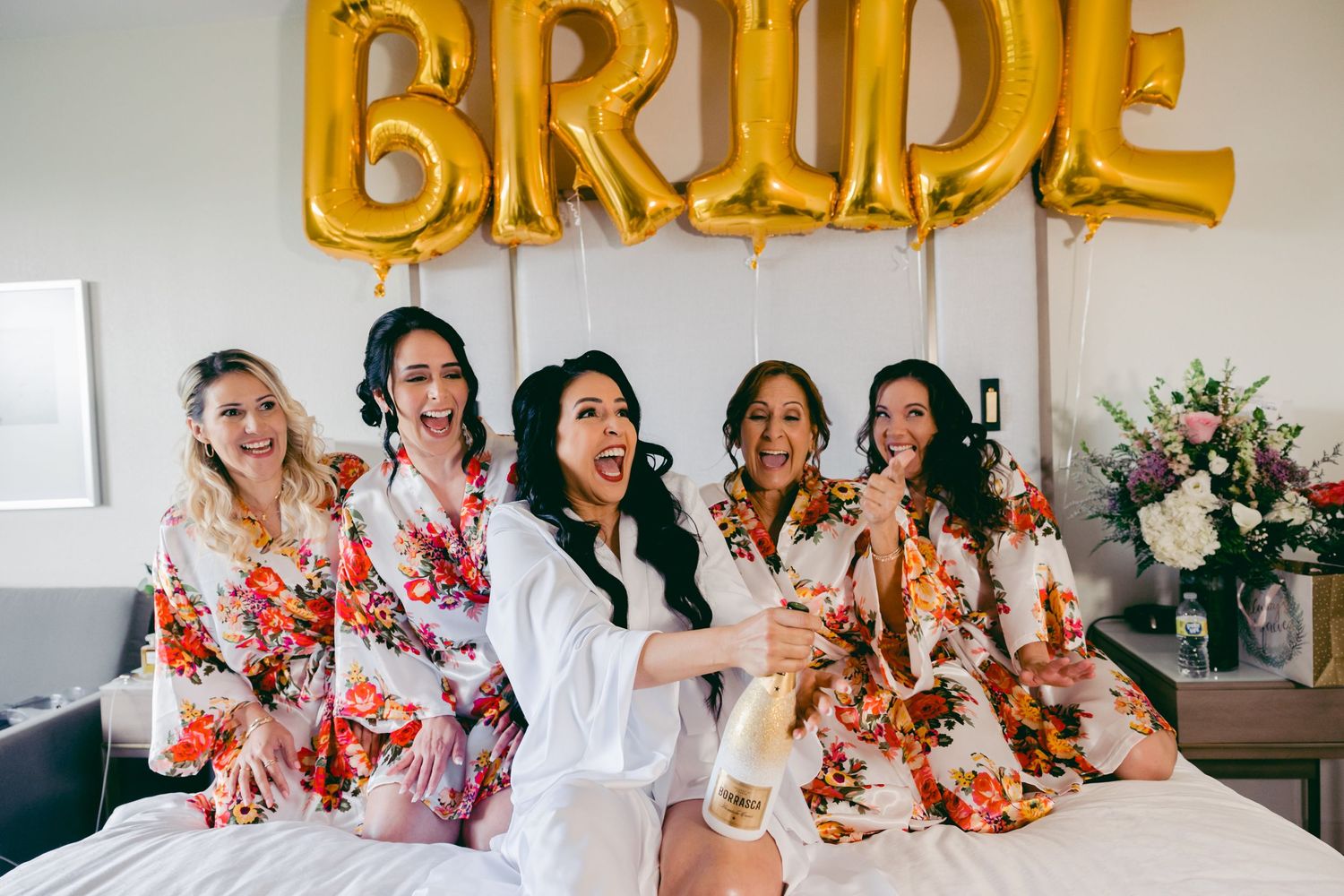 Wedding party in floral robes posing together under gold BRIDE balloon decoration.