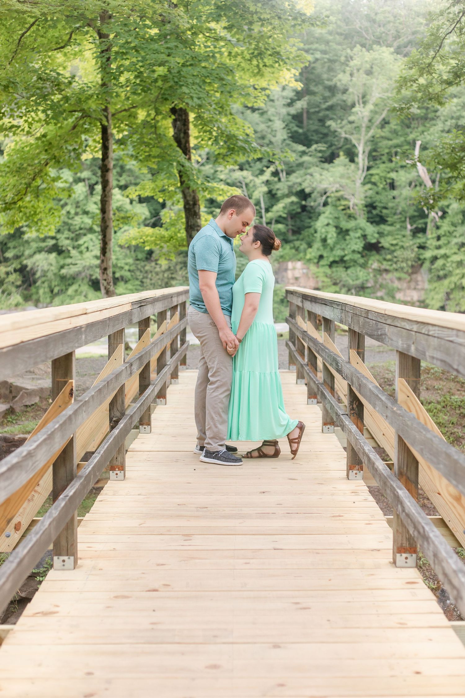 A couple shares a romantic moment on a wooden bridge surrounded by lush green forest.