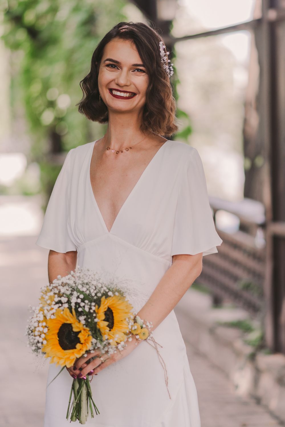 Bride in white dress holds sunflower bouquet while smiling on garden pathway.