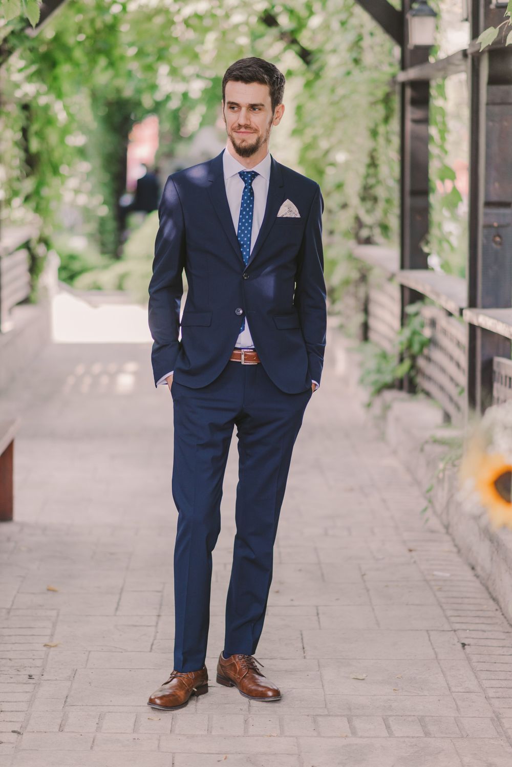 A groom in a navy blue suit and brown shoes stands on a stone pathway.