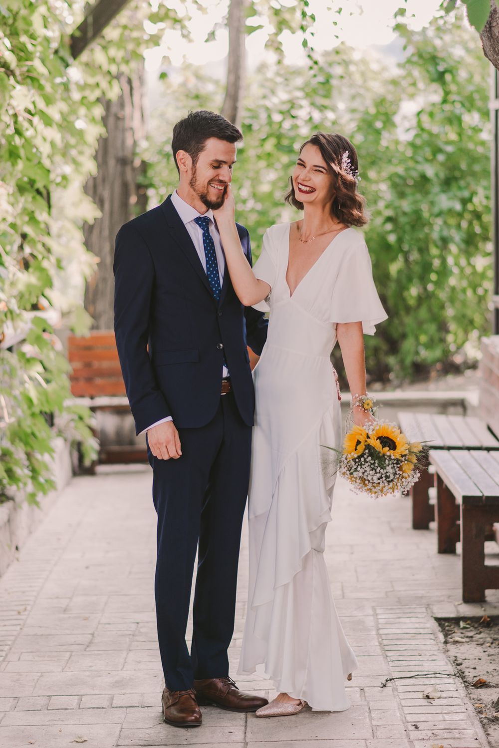Couple in wedding attire exchange glances while holding sunflower bouquet on garden path.