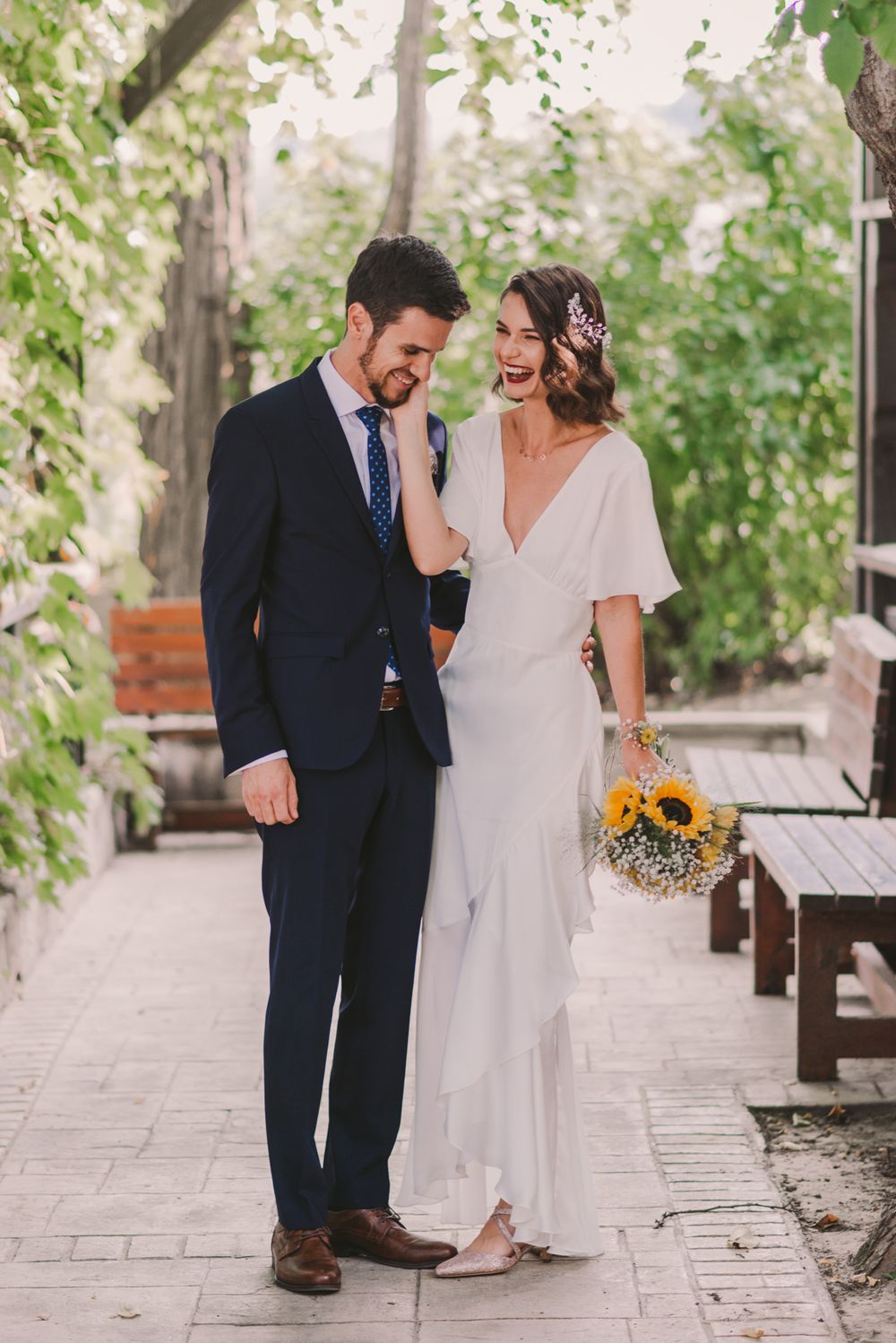 Newlyweds share joyful moment on stone pathway surrounded by green foliage.