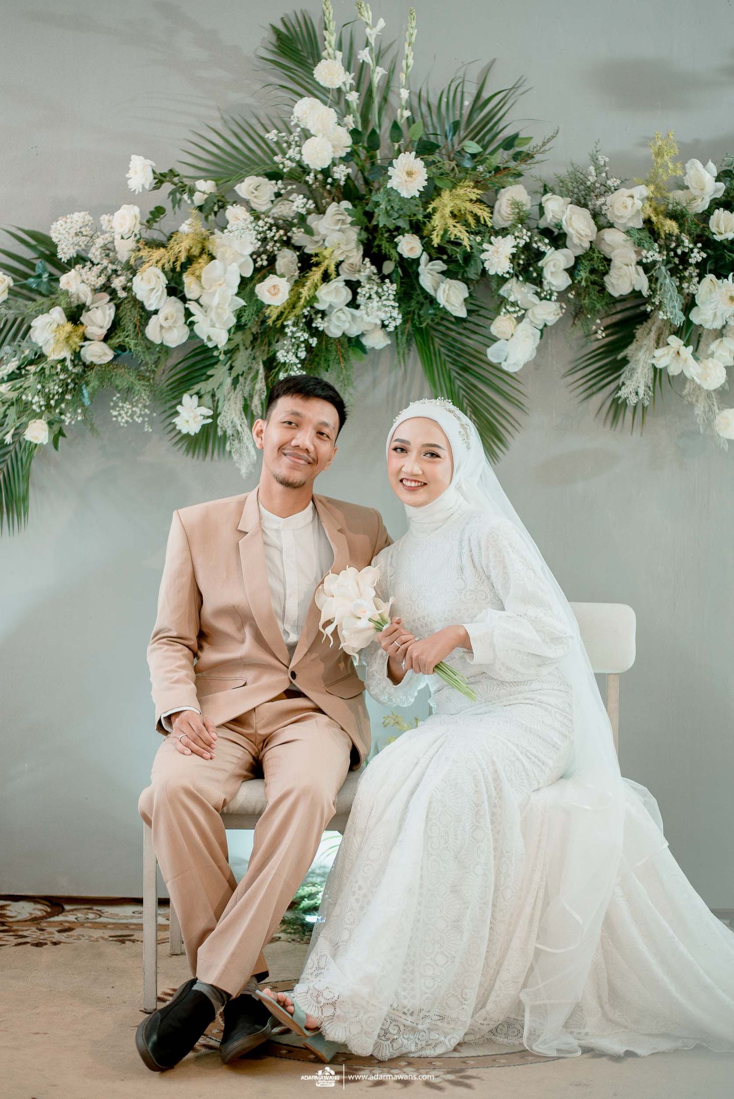 Wedding couple sits beneath a luxurious floral arch with white flowers and palm leaves against a grey backdrop.