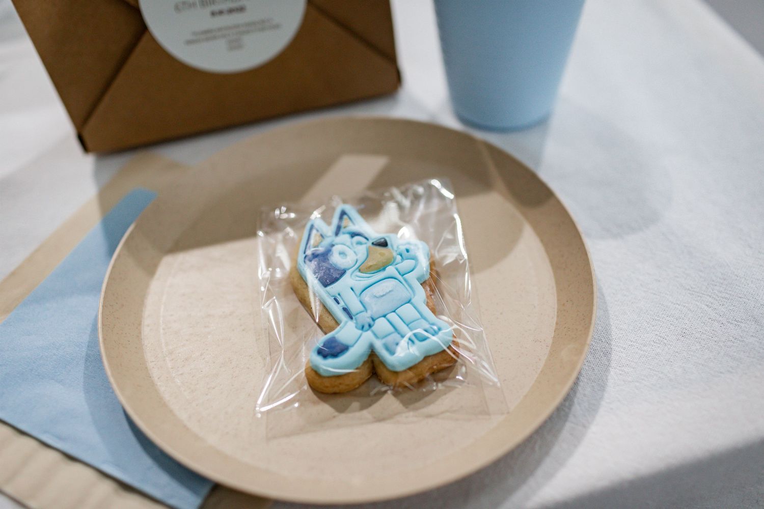 Blue decorative cookie wrapped in plastic on a kraft paper plate with party supplies.