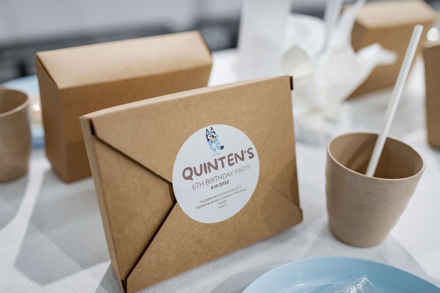 Kraft paper party favor boxes with 'Quinten's' stickers next to eco-friendly cups.