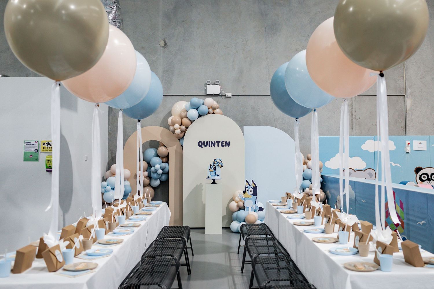Party setup with blue and beige balloons lining long tables in an arch-decorated event space.