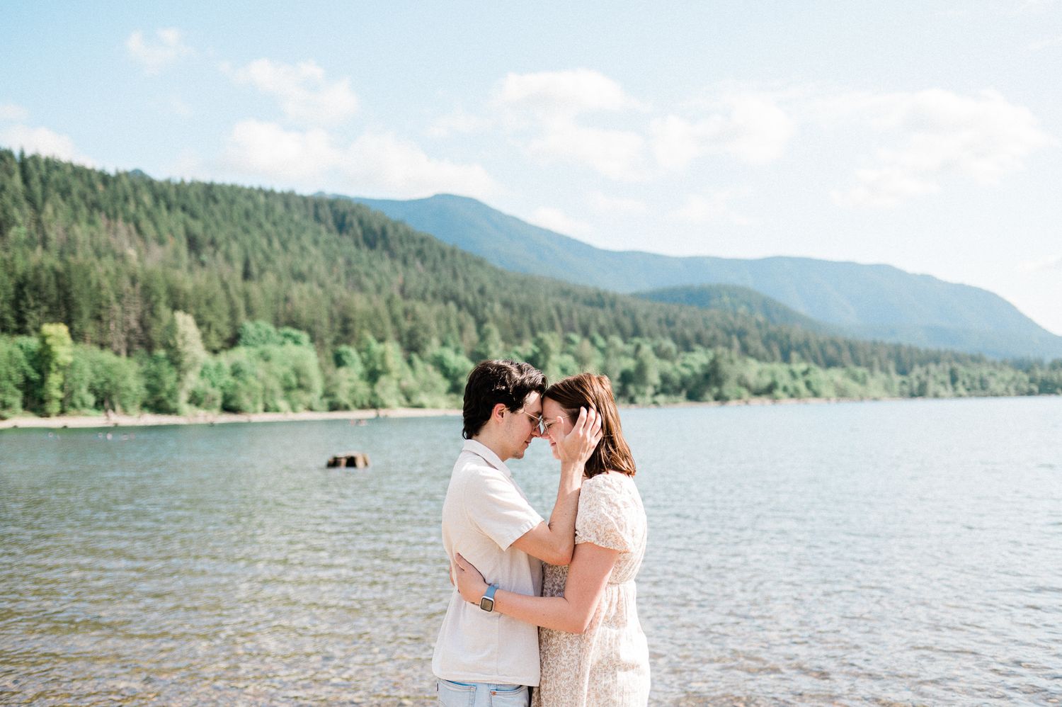 A couple shares a romantic moment on a scenic mountain lake shoreline with evergreen trees in the background.