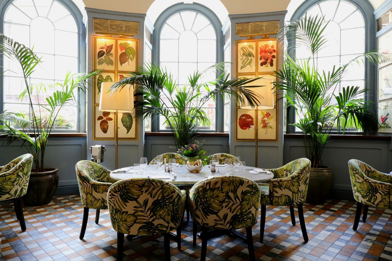 Elegant dining room with arched windows, tropical plants, and upholstered chairs featuring ornate artwork on the walls.
