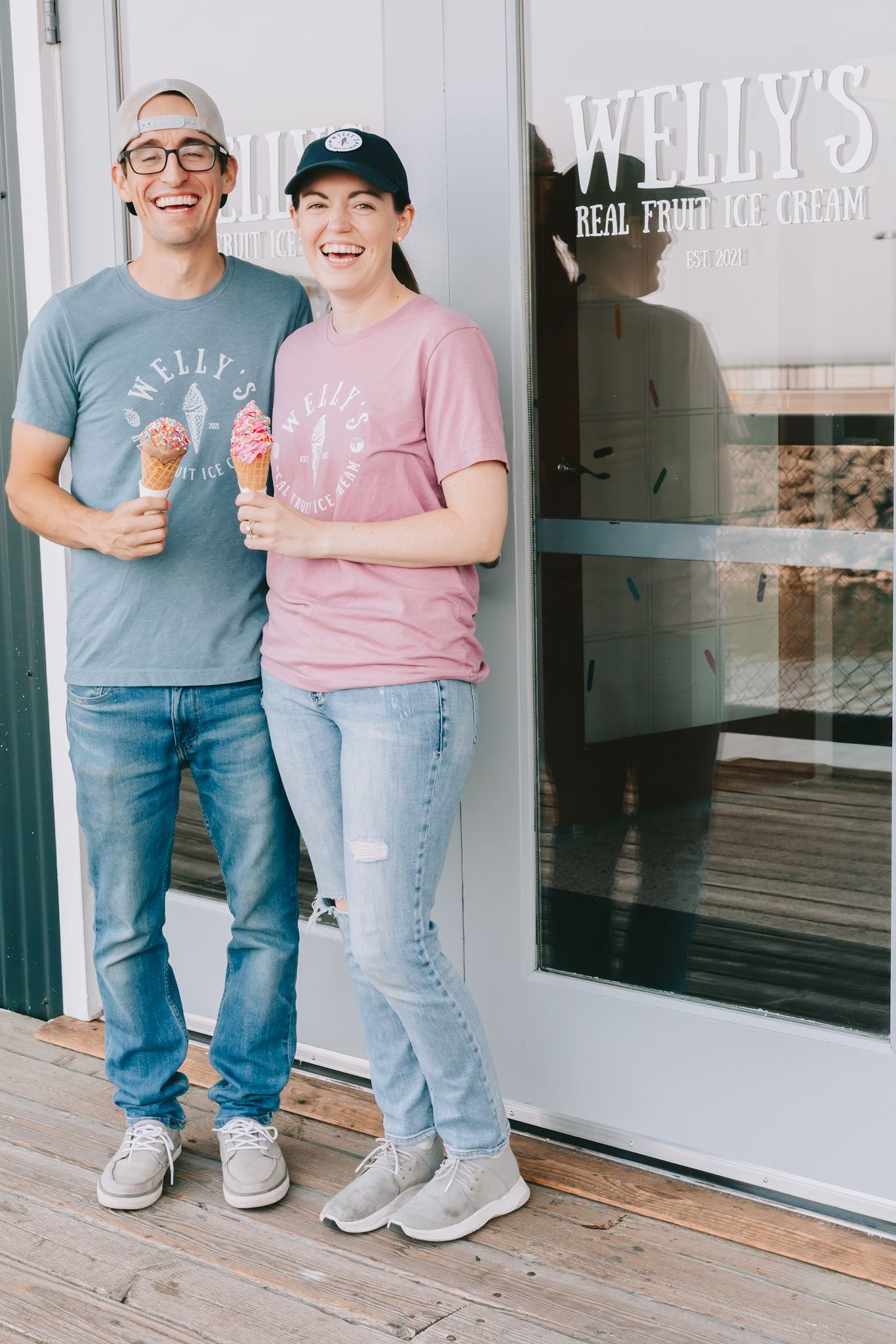 A couple wearing Welly’s Real Fruit Ice Cream t shirts stand with ice cream cones infront of a door that says welly’s real fruit ice cream.