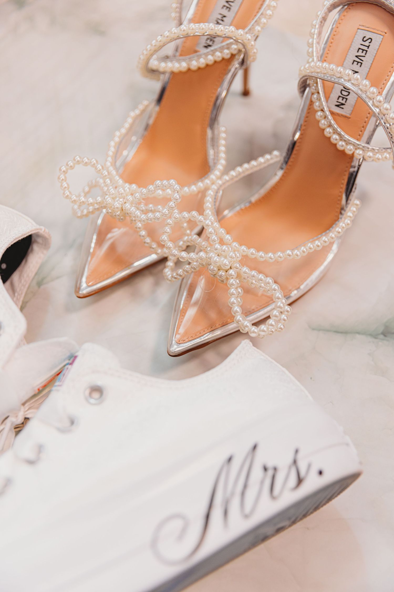 Steve Madden pearl strappy heels next to white Mrs. custom bridal sneakers on marble surface.