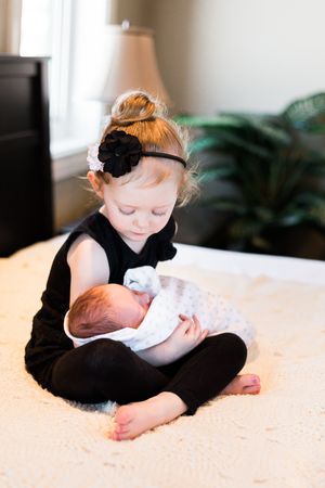 Young toddler girl in black outfit gently holding a swaddled newborn baby while sitting on a bed.
