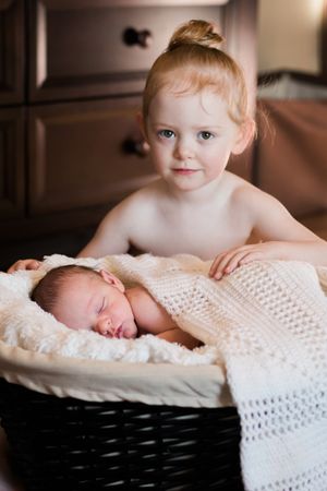 An adorable toddler girl with a bun hairstyle stands behind a basket holding a sleeping newborn.