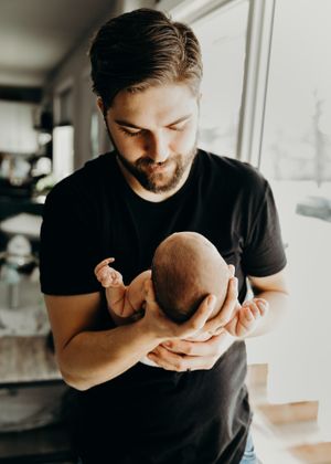 Bearded father in black shirt tenderly holding his newborn baby near a bright window.