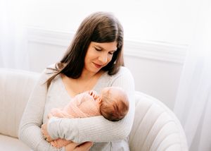 Brunette mother in grey sweater holding her sleeping swaddled newborn in a bright white room.