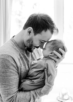 Black and white photo of father tenderly kissing newborn baby by window.