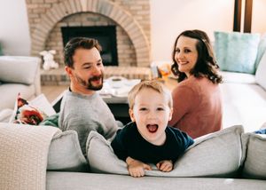 Happy family with toddler laughing on couch in front of brick fireplace.