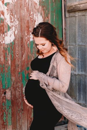 Pregnant woman in black dress with lace wrap cradling baby bump near rustic wall.
