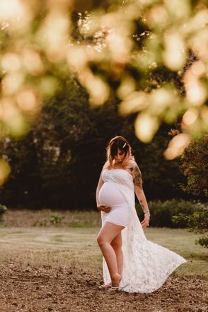 Pregnant woman in flowing white lace gown posing in sunlit garden at golden hour.