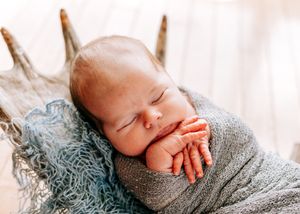 Peaceful newborn baby sleeping wrapped in a gray swaddle on a rustic wooden prop.