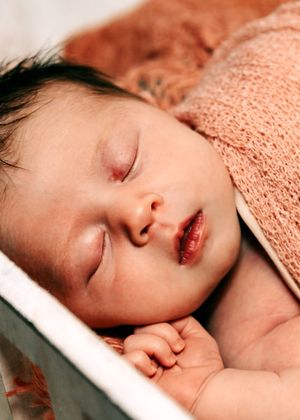 Sleeping newborn baby with dark hair resting peacefully under a peach knit blanket.