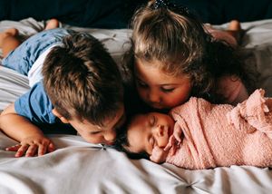 Two young siblings lying on a bed giving gentle kisses to their pink-swaddled newborn.
